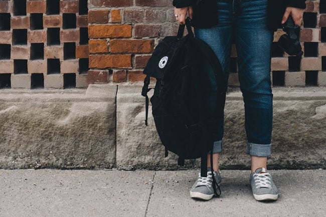 A person stands on a sidewalk holding a black backpack