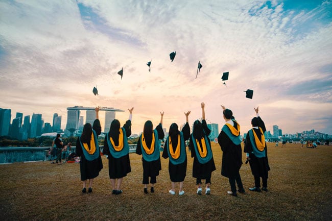 A group of students stands on a hillside tossing their graduation caps into the air