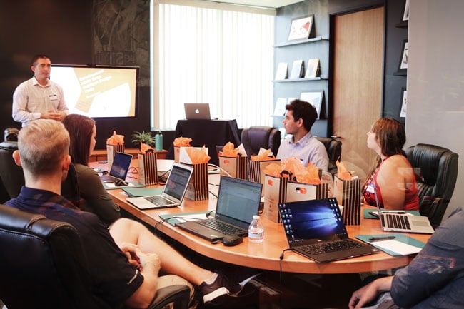 Several people are seated around a large table filled with laptop computers, while one person stands at the head of the room