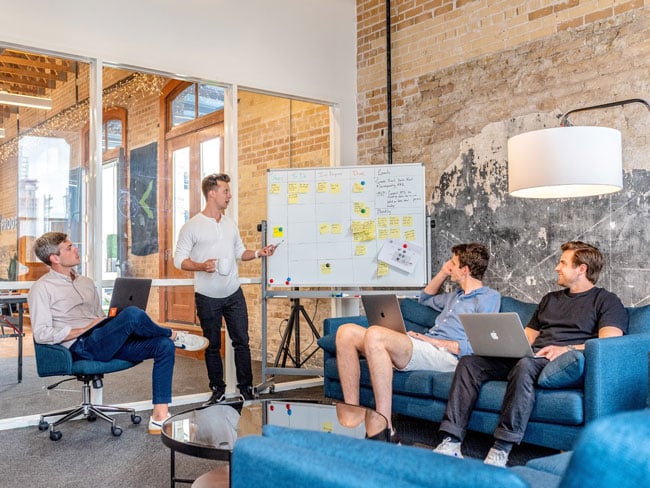 Four men are in an office while one stands at a whiteboard covered in yellow notepaper